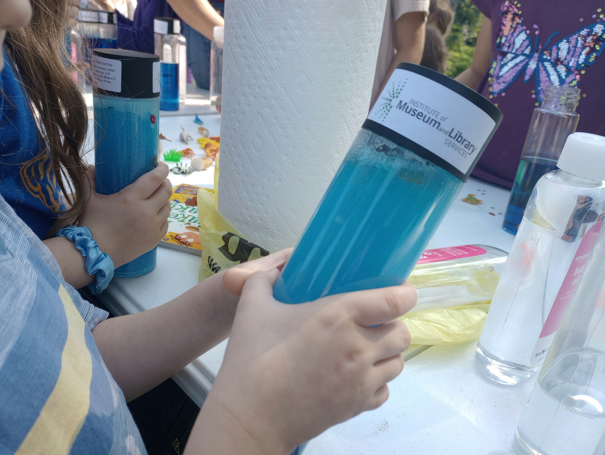 Children holding blue liquid-filled bottles at a craft table with paper towels and colorful items.