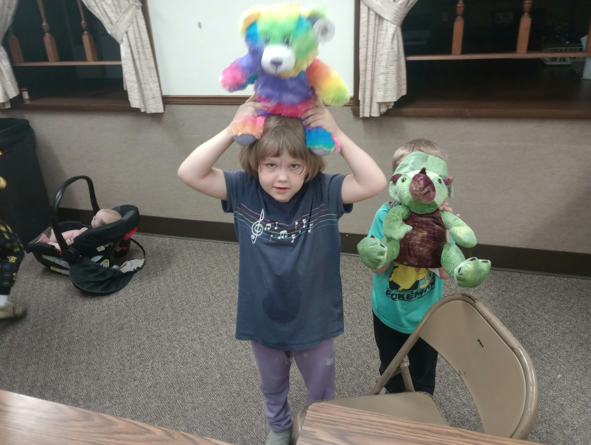 Children holding stuffed toys, one with a colorful teddy bear, another with a green dinosaur plush, indoors near a baby carrier.