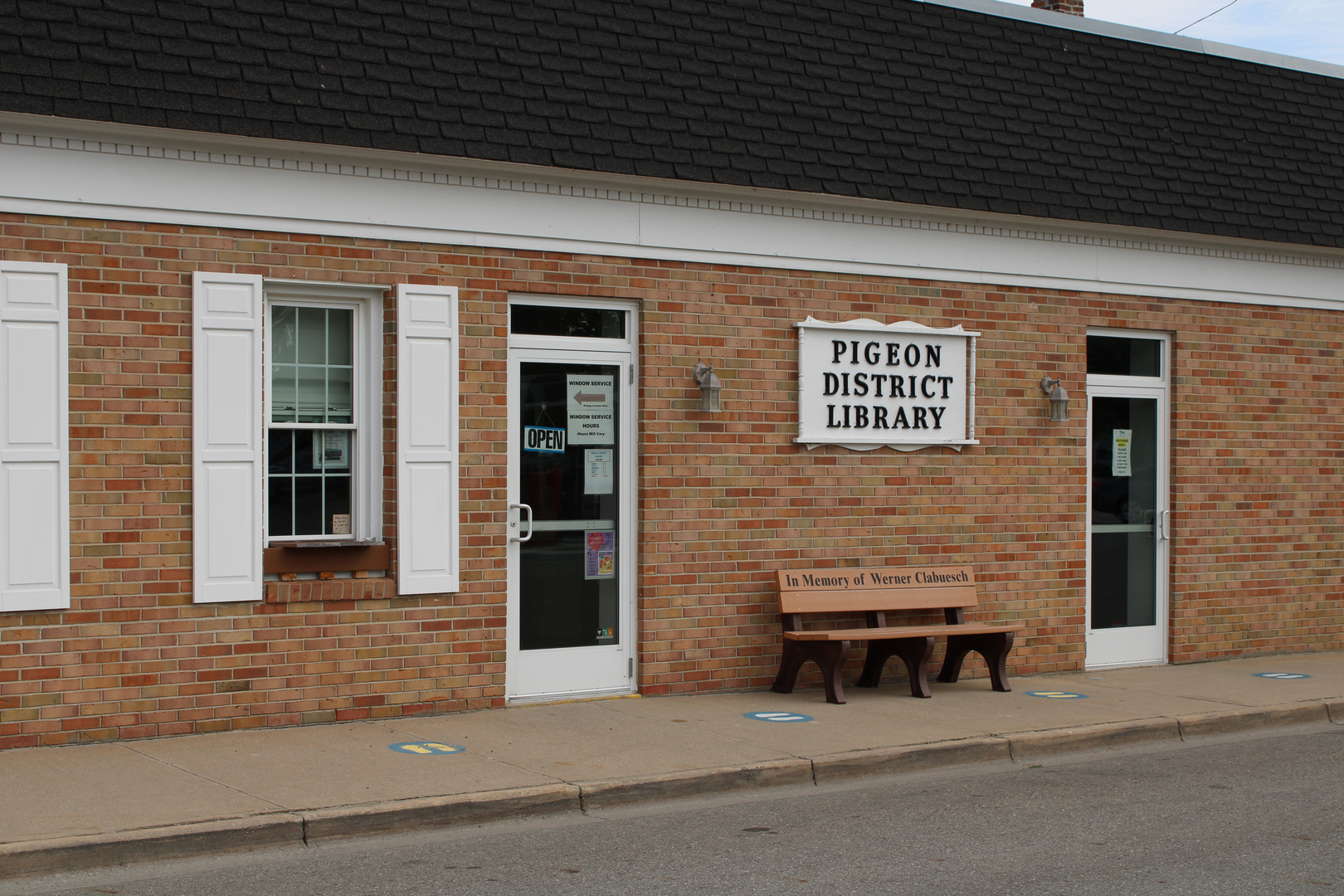 The image shows the Pigeon District Library's brick exterior, featuring an open sign, white shutters, and a memorial bench.