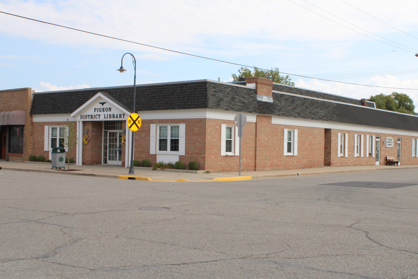 The image shows the Pigeon District Library, a single-story brick building with a sign and some greenery.