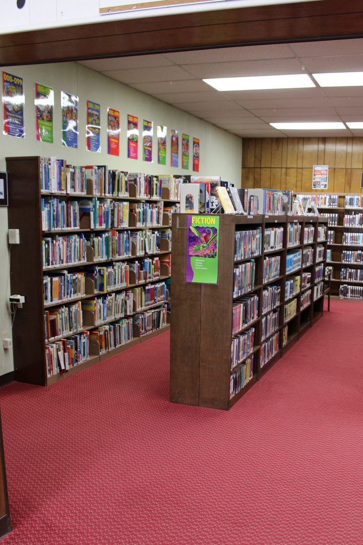 The image shows a library with bookshelves filled with books, a fiction section highlighted, and a red carpet floor.