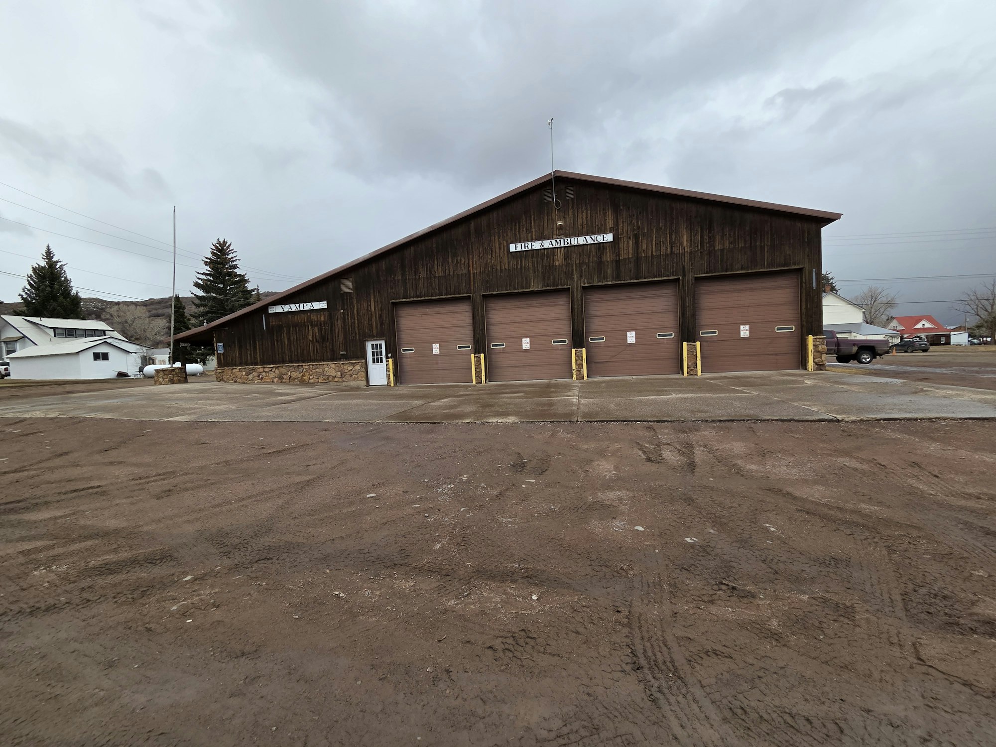The image shows a wooden building with multiple garage doors, likely serving as a fire and ambulance station, against a cloudy sky.