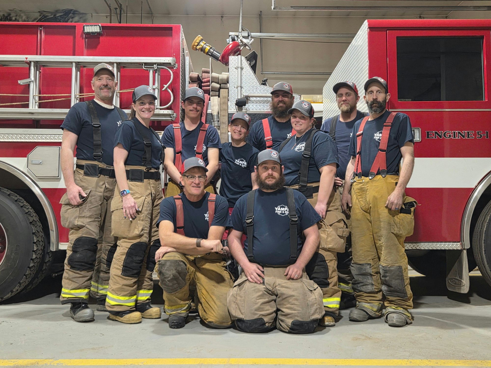 A group of firefighters poses together in front of their fire truck, all wearing uniforms and gear.