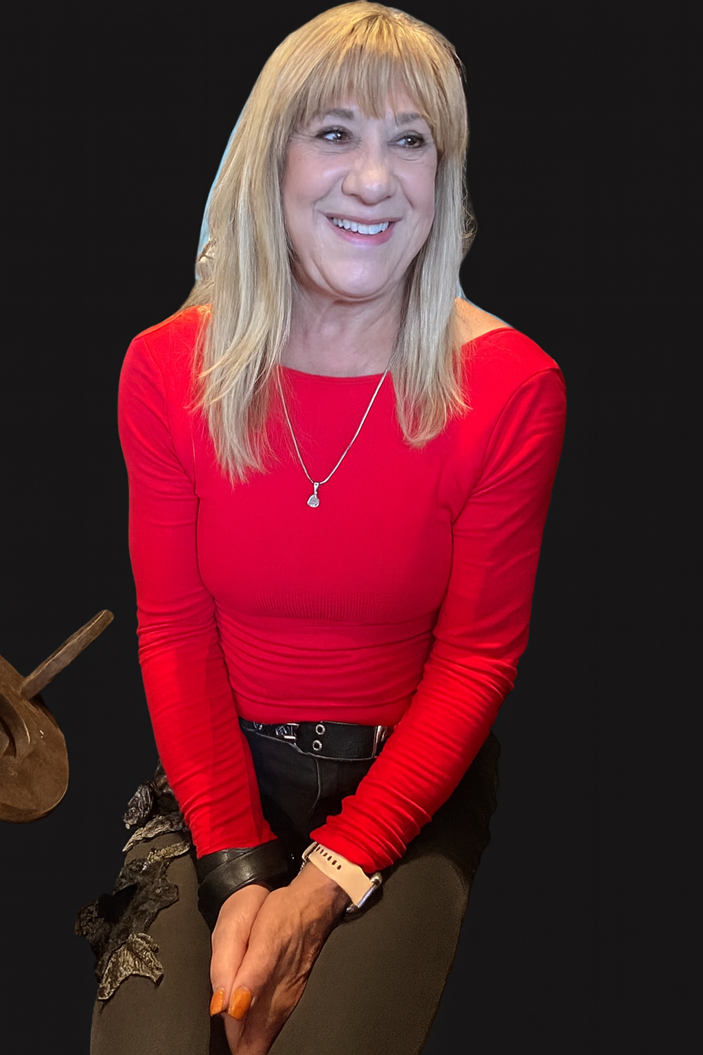 A person with long hair wearing a red top, black pants, and a watch, smiling against a dark background.