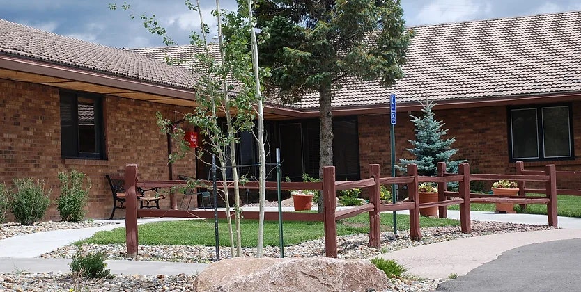 A building with a brick facade and sloped roof, surrounded by a landscaped area with young trees, a bench, and a pathway.