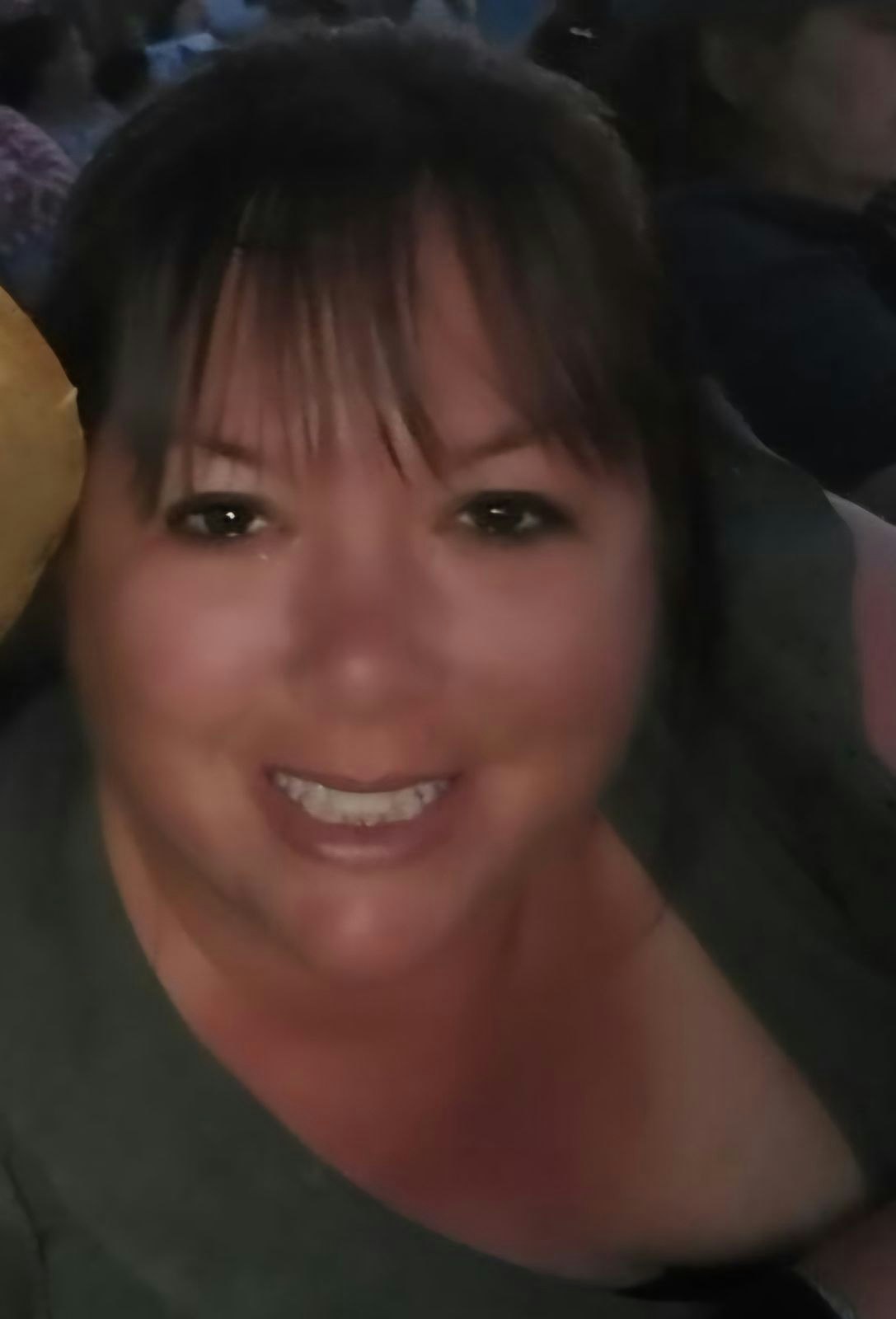 A smiling woman's selfie, possibly at an event with a crowd in the background.