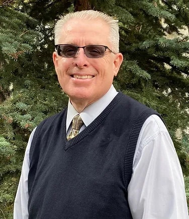 A smiling man in sunglasses, wearing a tie, shirt, and sweater vest, with trees in the background.