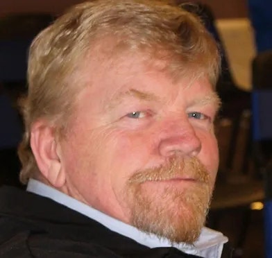 A man with a beard and mustache, wearing a black jacket, looking slightly to the side.