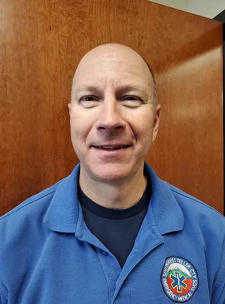 A smiling person wearing a blue shirt with an emergency medical service badge in front of a wooden background.