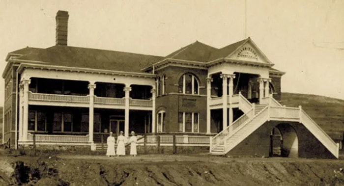 Historic building with verandas and three figures in white attire, sepia-toned photo.