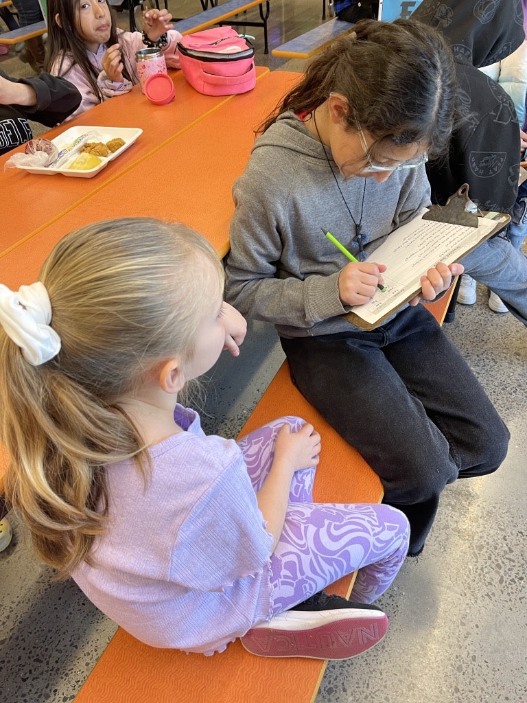 Two children sitting on an orange mat; the older one is writing on a clipboard while a younger child looks on.