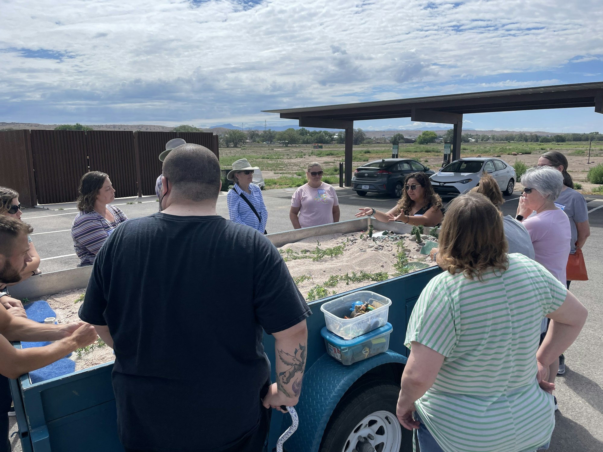 Group of people around a table with plants, in an outdoor setting with vehicles and a cloudy sky.