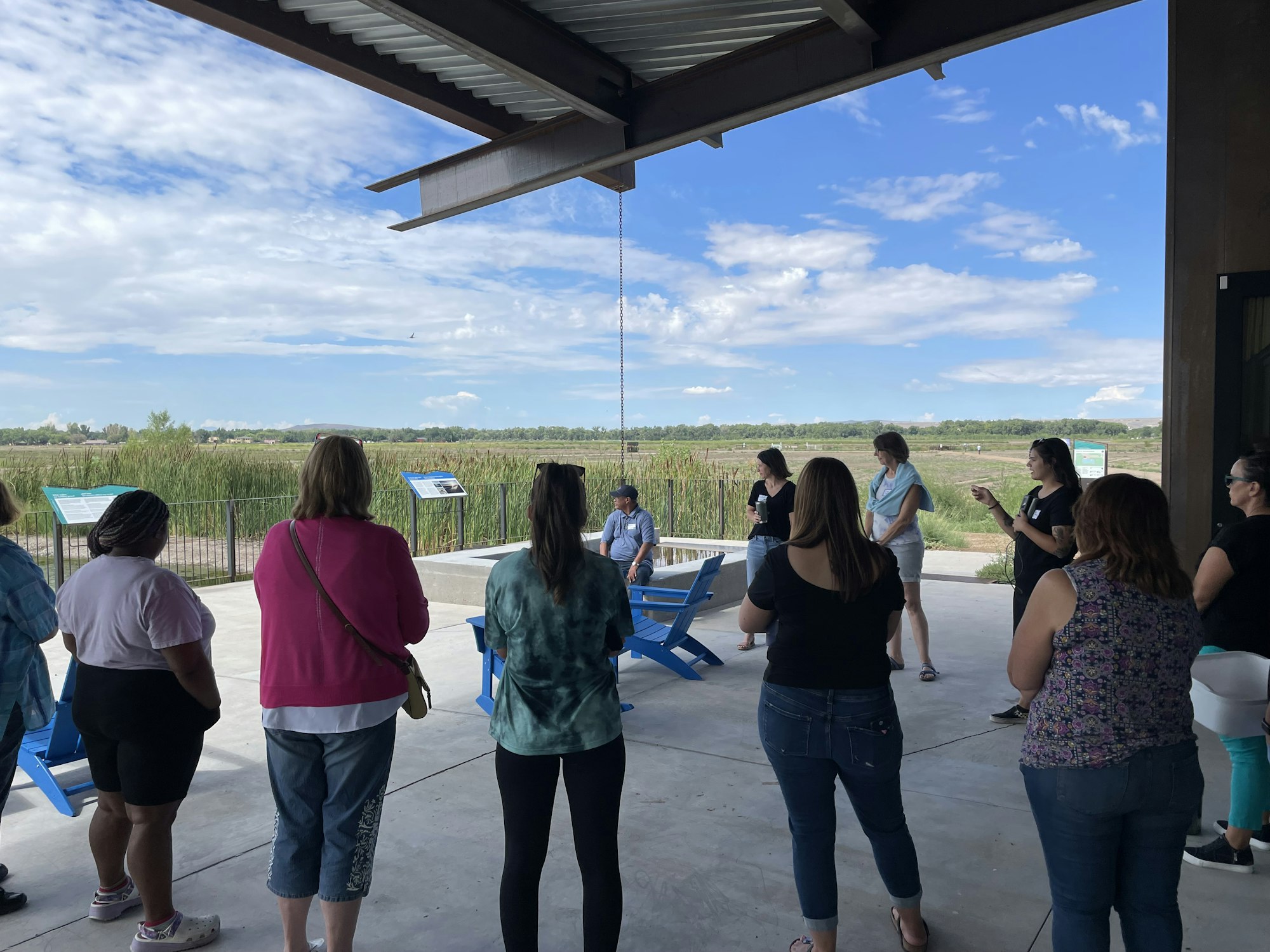 A group of people gathered on a patio, possibly attending an outdoor event or meeting, with a scenic nature backdrop.