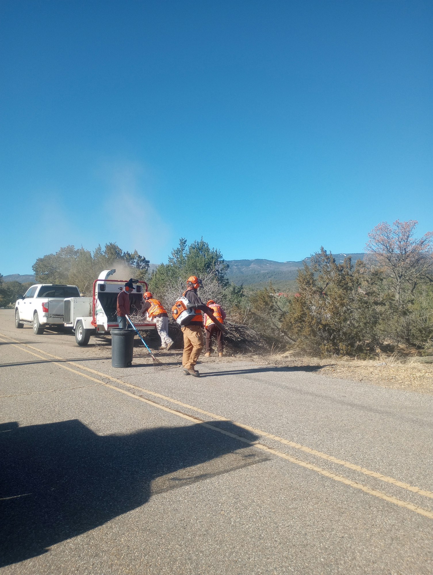 Workers in orange vests on a roadside near trucks and equipment, with clear blue sky above.