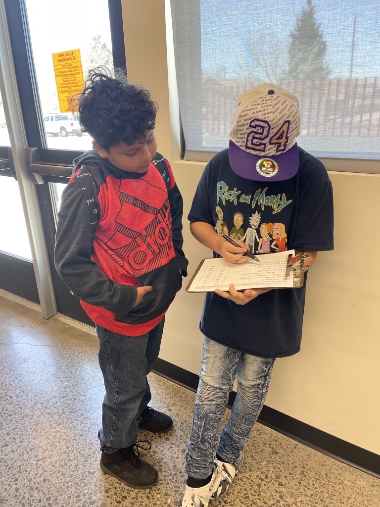 Two boys, one writing, indoors, with a window behind them.