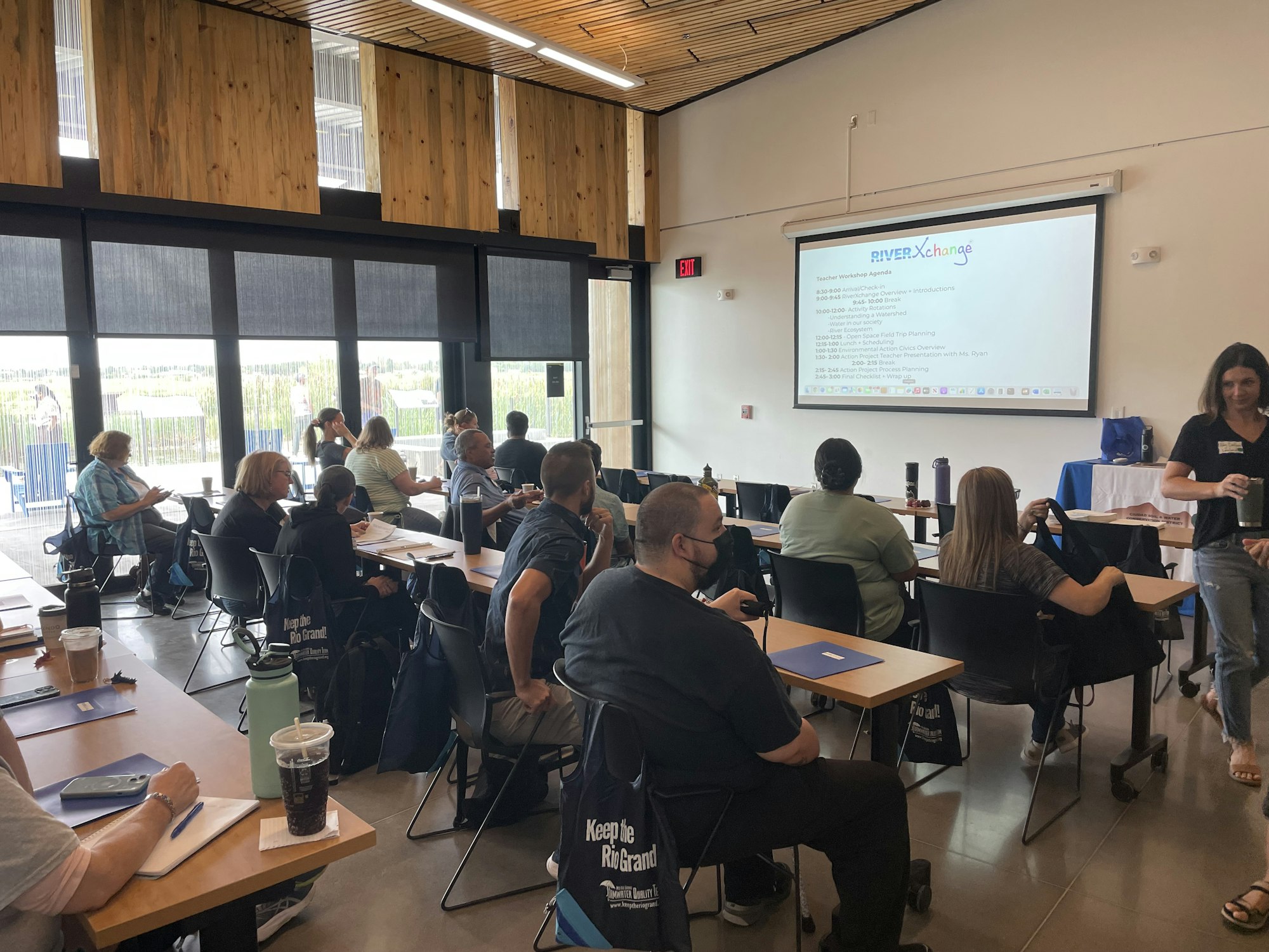 A group of people in a meeting or workshop with a presentation screen titled "RIVER Xchange" in a modern room with large windows.