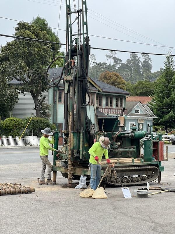 Two workers in safety gear operate drilling equipment on a street.