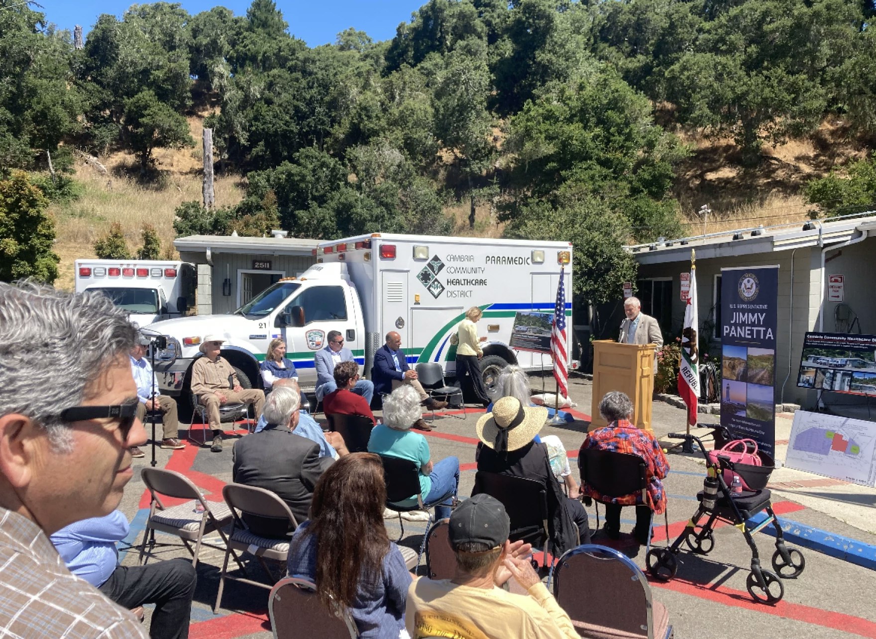 An outdoor event with a speaker at a podium, an audience seated, and an ambulance in the background.