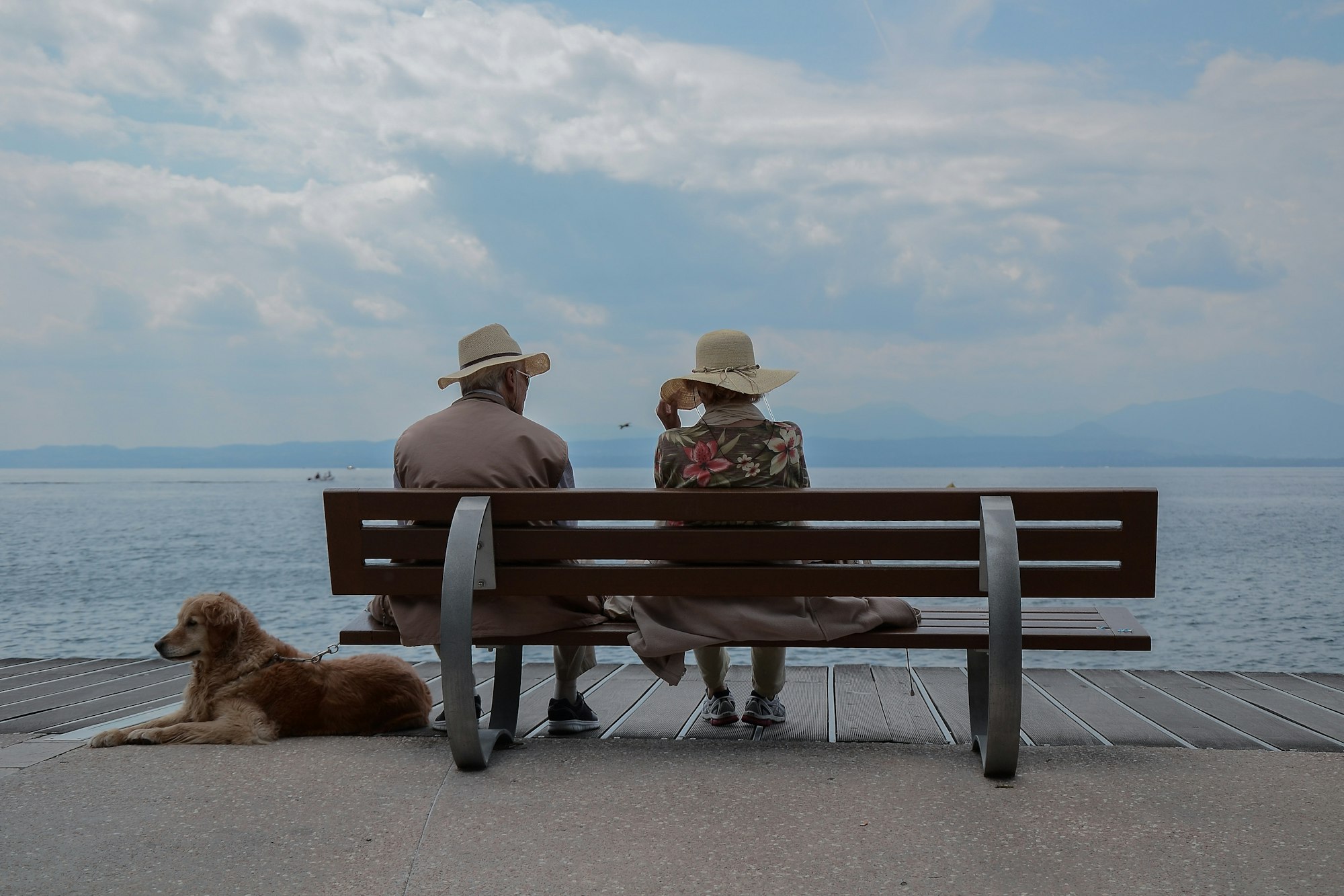 Two people with hats sit on a bench by the sea with a dog resting beside them.