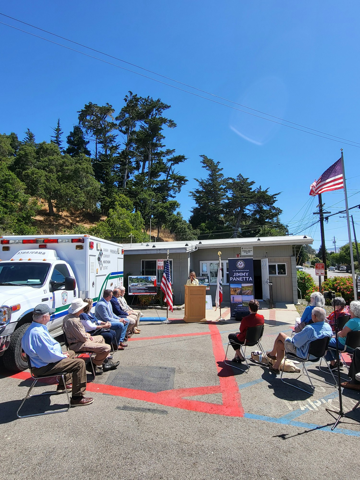 Outdoor gathering with American flags, people sitting on chairs, and ambulance in the background. (Note: Image is rotated)