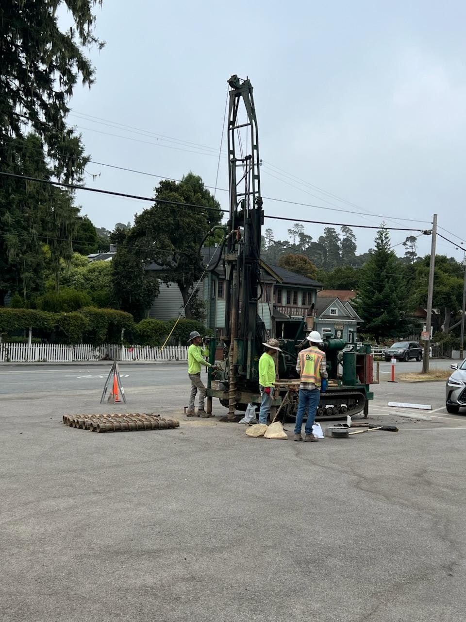 Construction workers operate drilling equipment in a parking lot, with trees and houses in the background.