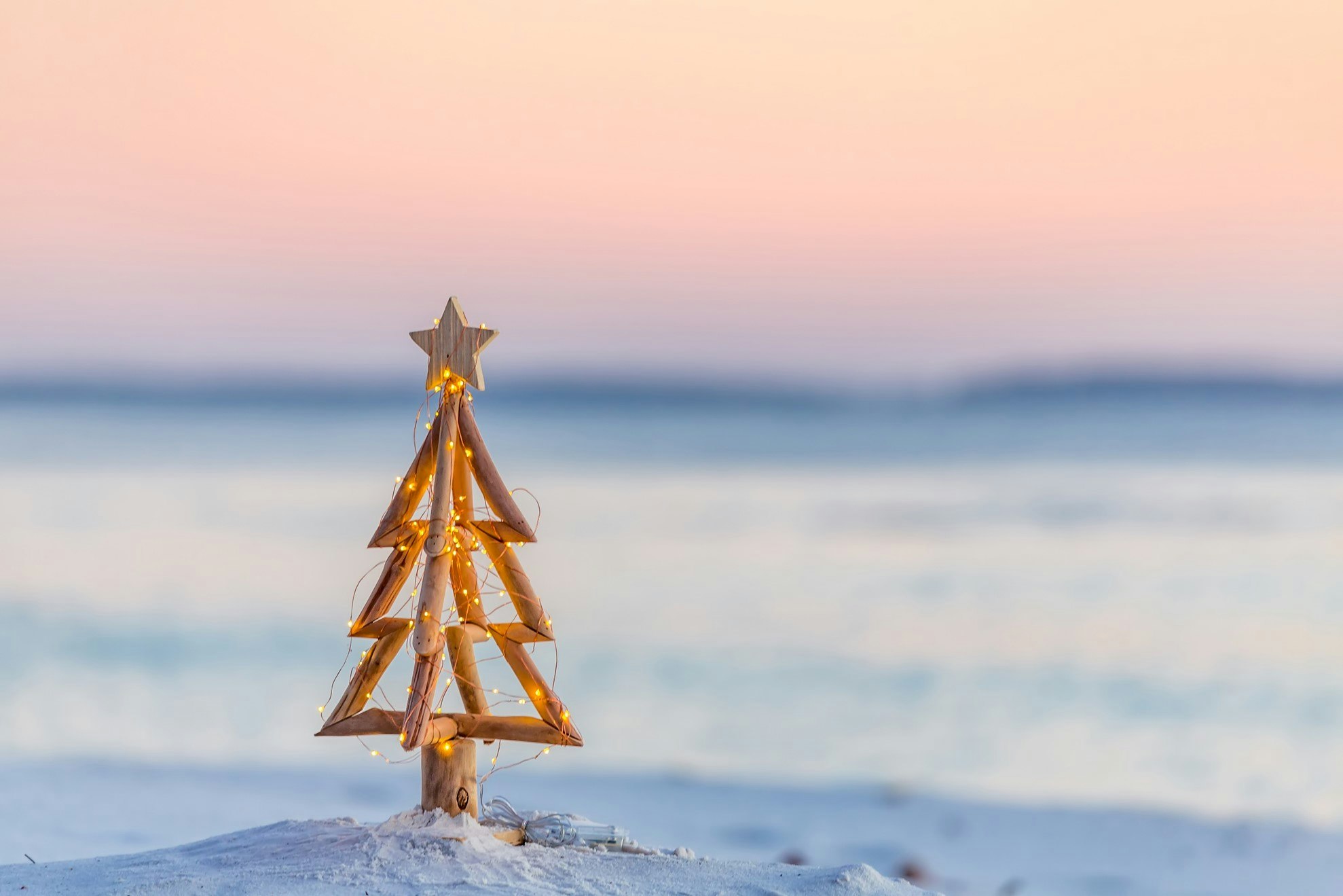 A small wooden Christmas tree with lights on a sandy beach at sunset.