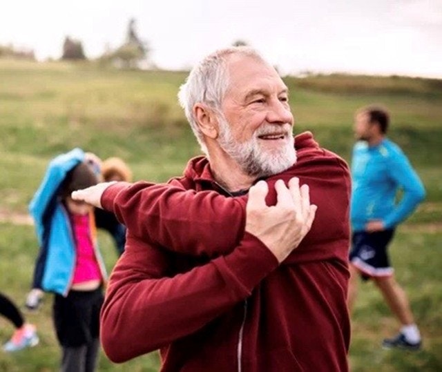 An elderly man stretches outdoors with others in the background, wearing a red jacket.