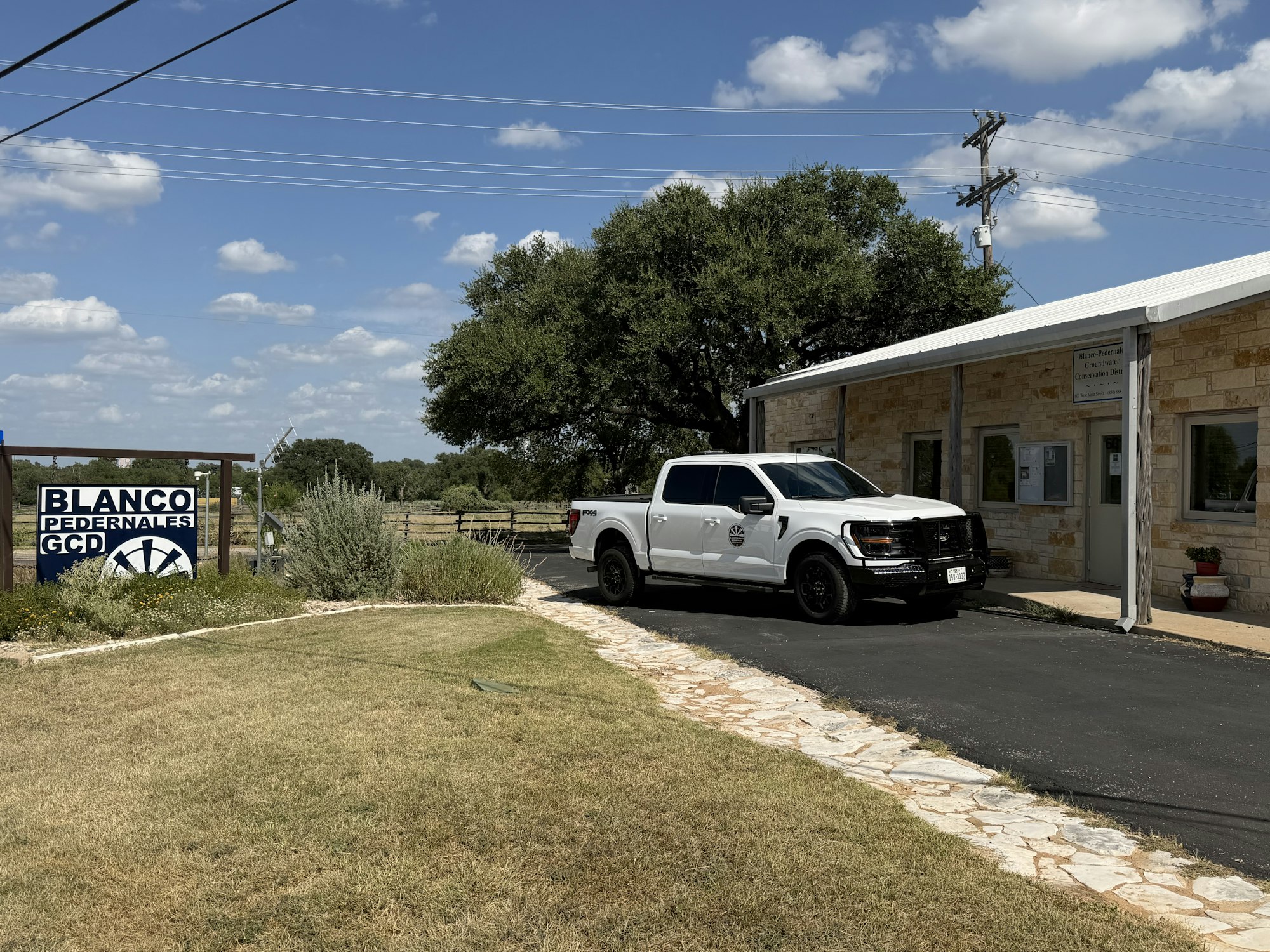 The image shows a white pickup truck parked by the Blanco-Pedernales Groundwater Conservation District office in a rural setting.