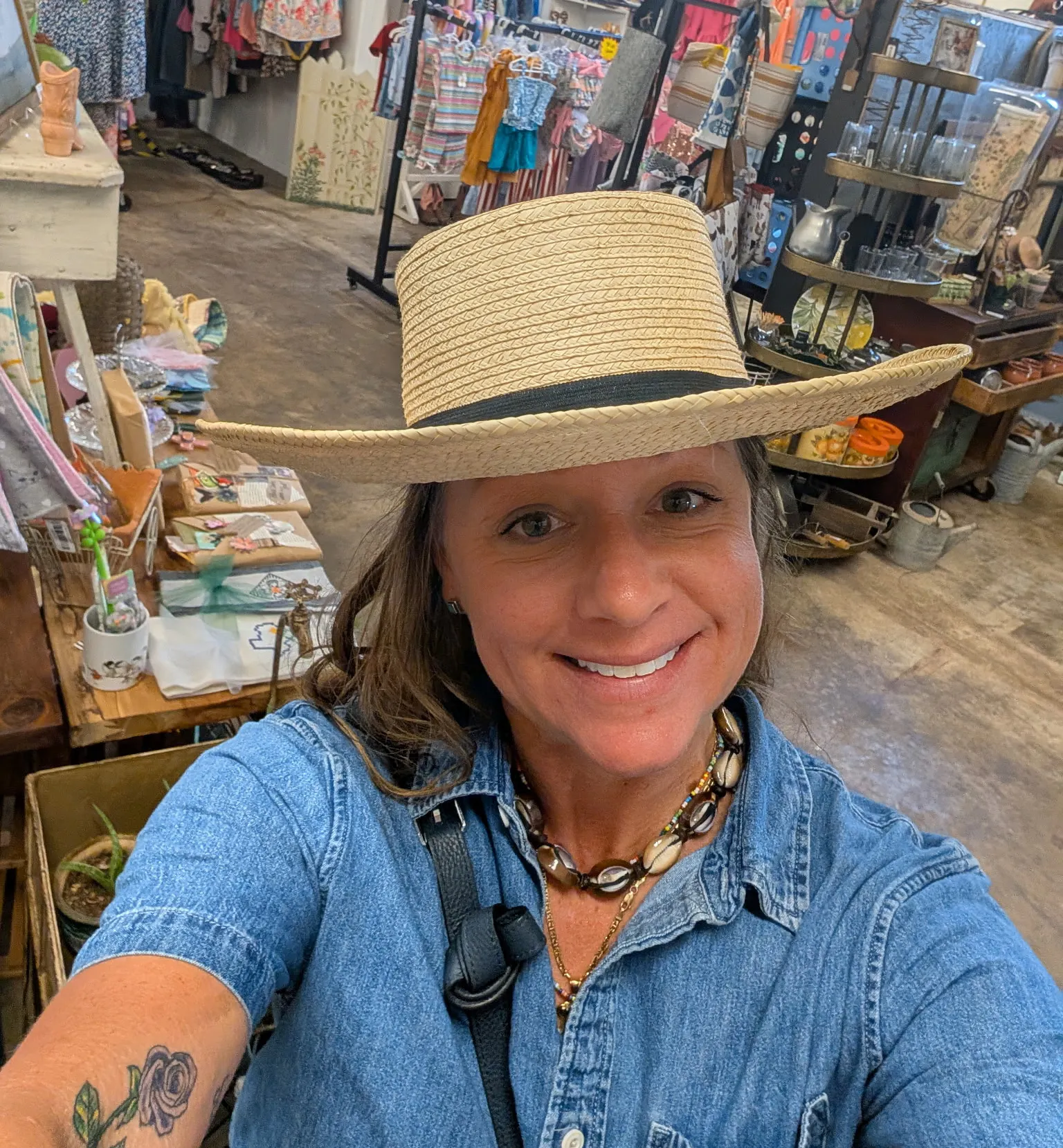A smiling person in a straw hat takes a selfie in a colorful boutique filled with clothing and accessories.