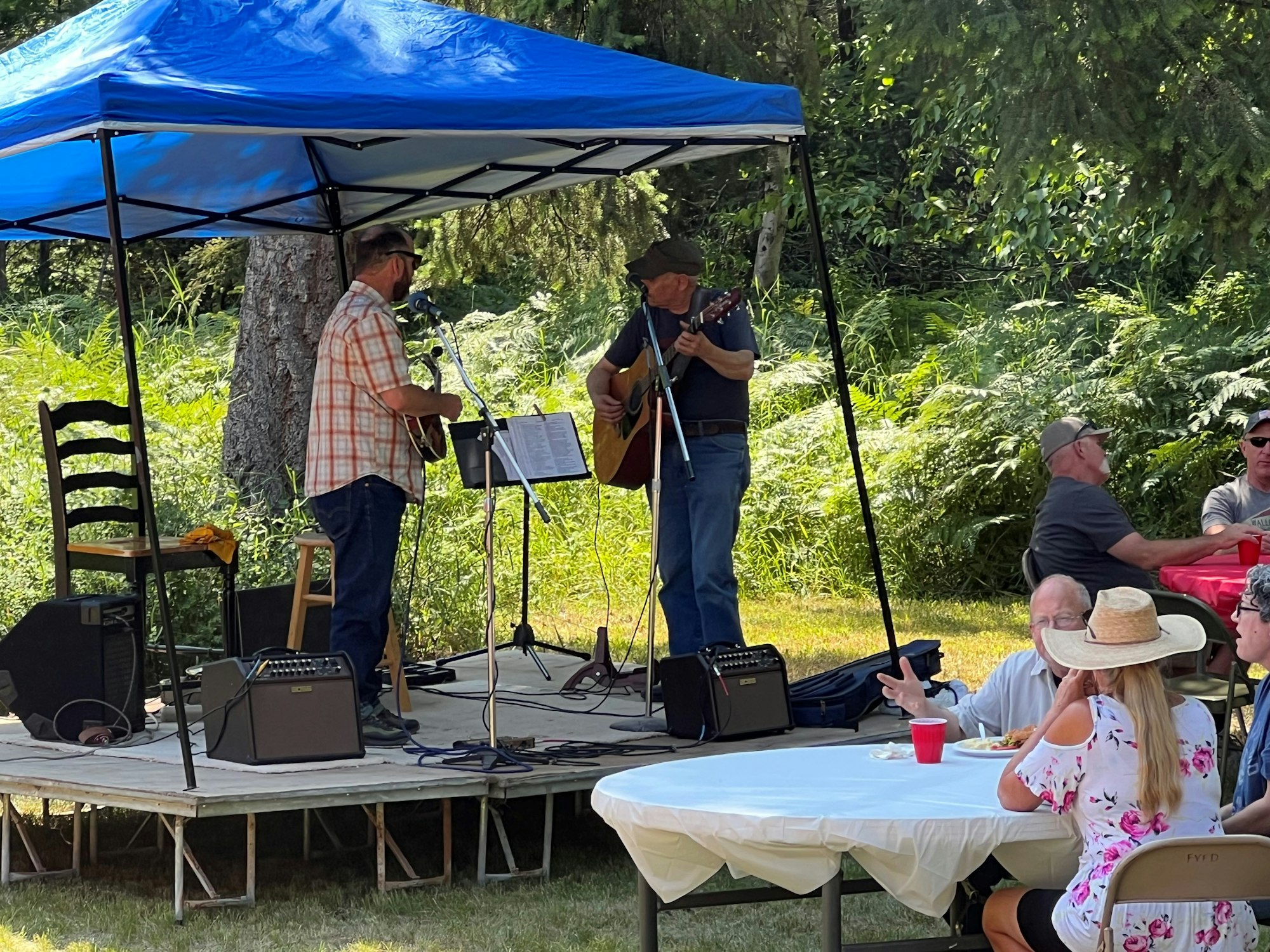 Two musicians playing guitars under a blue canopy with an audience seated at tables outdoors.