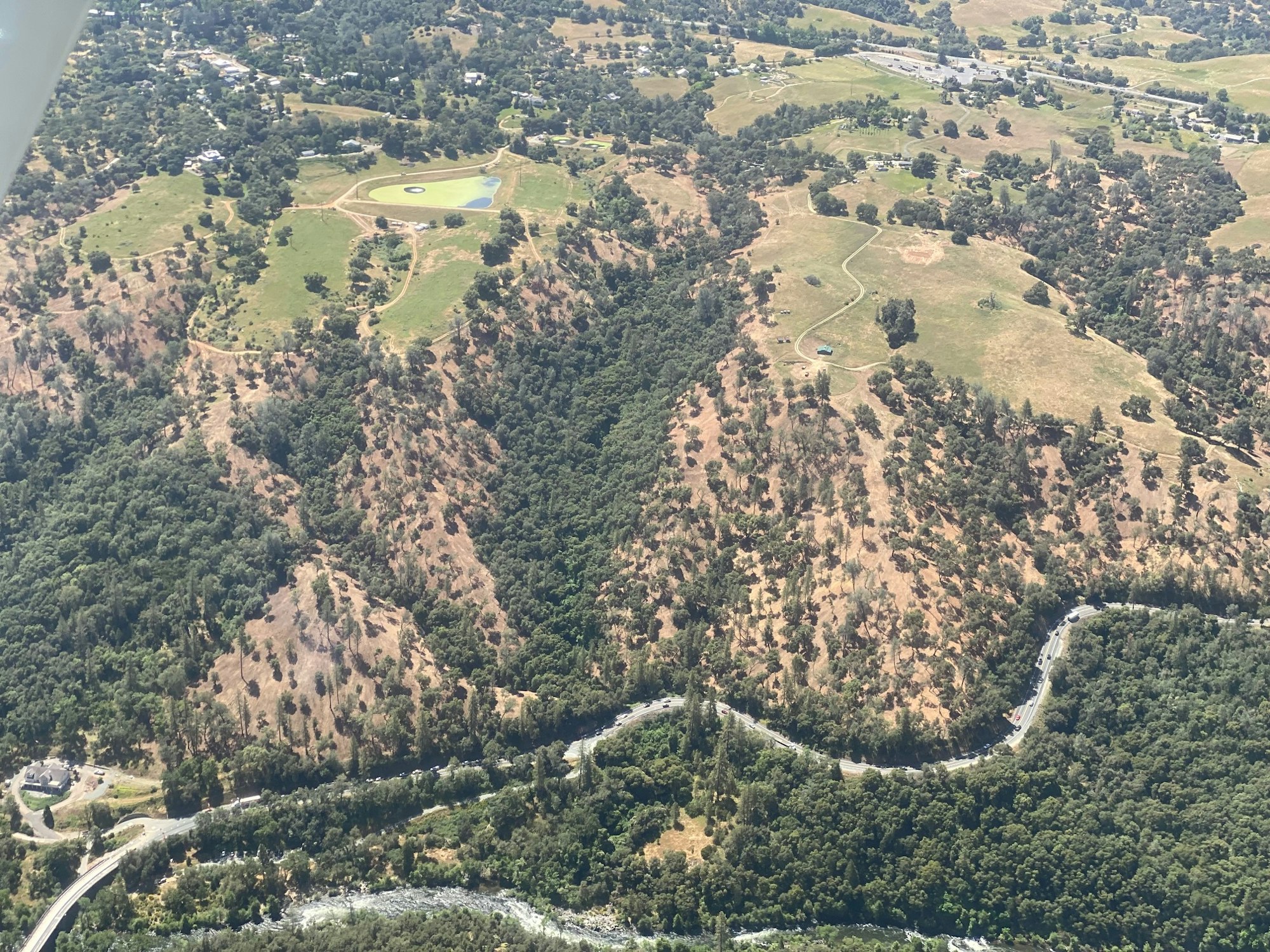 Aerial view of a hilly landscape, roads, trees, patches of grass, and a river.