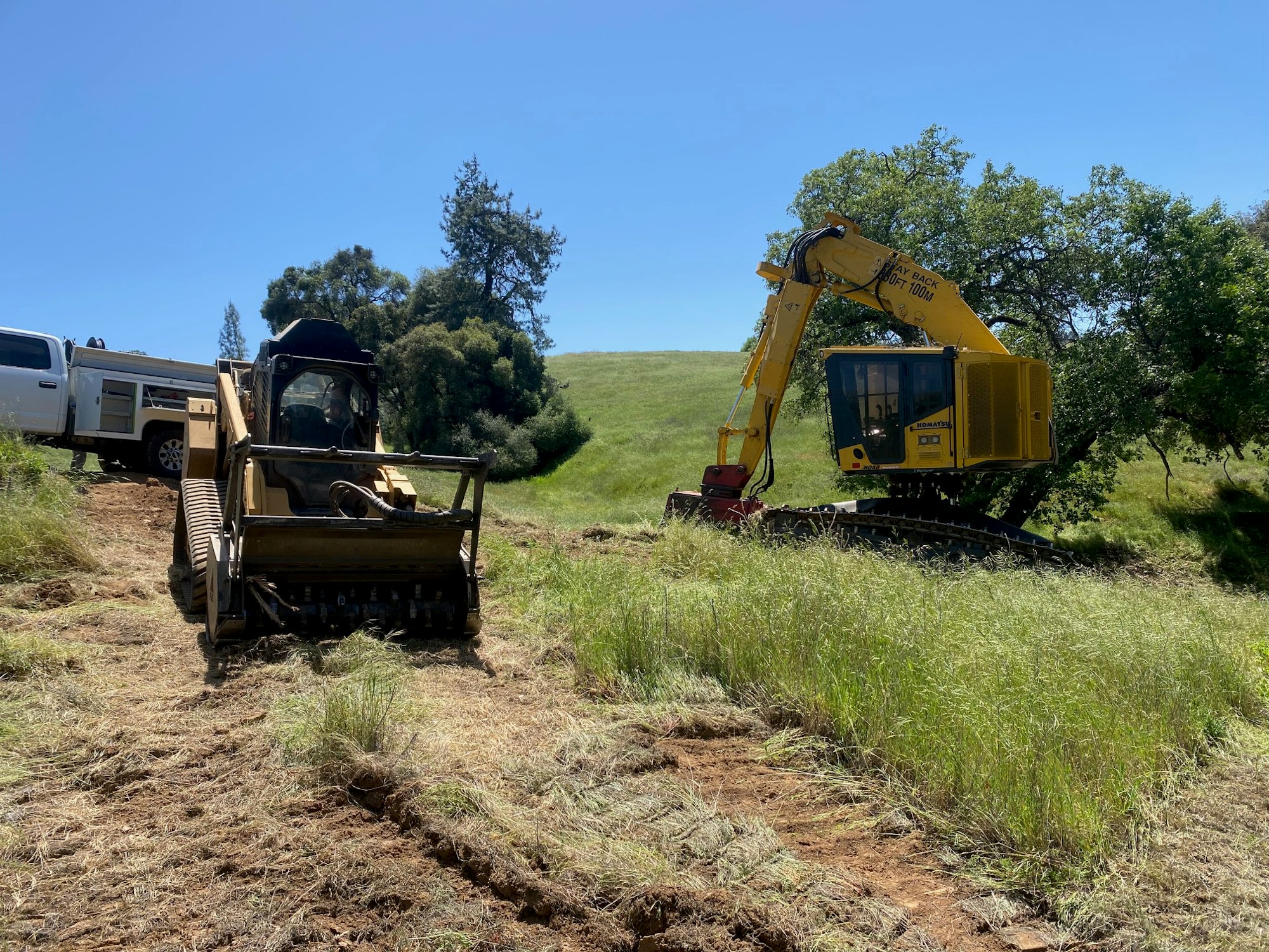 Heavy machinery (bulldozer and excavator) on a grassy field under a blue sky, possibly on a construction or land clearing site.