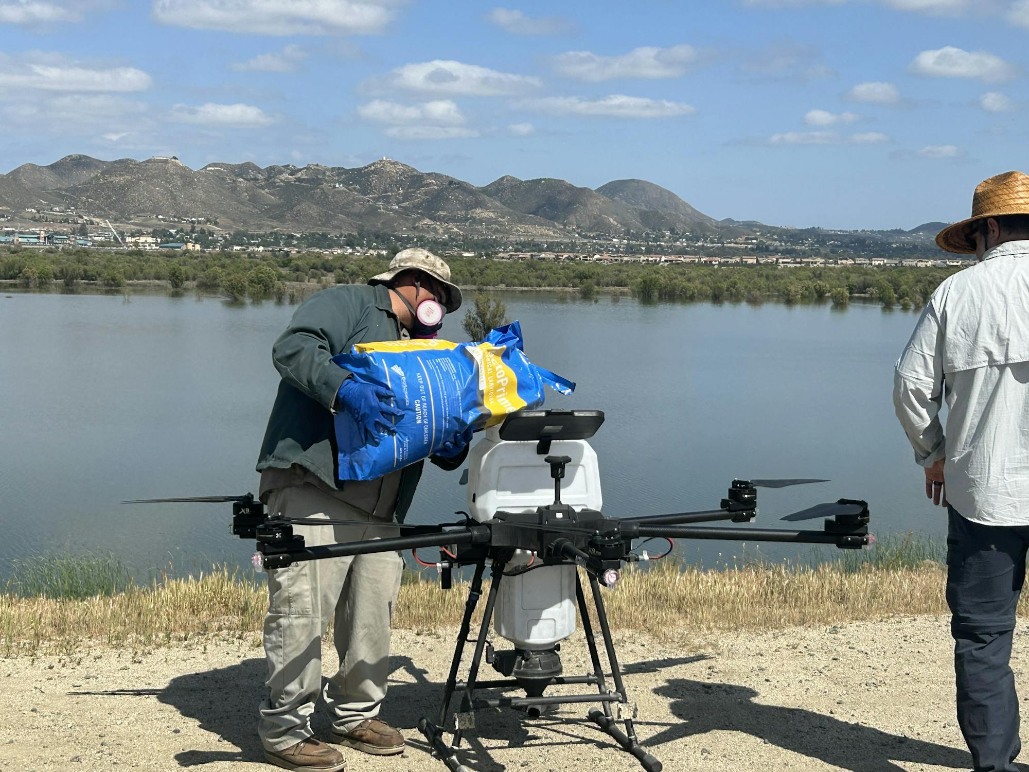 Two people by a lake; one is loading a drone with a blue and yellow bag under sunny skies.