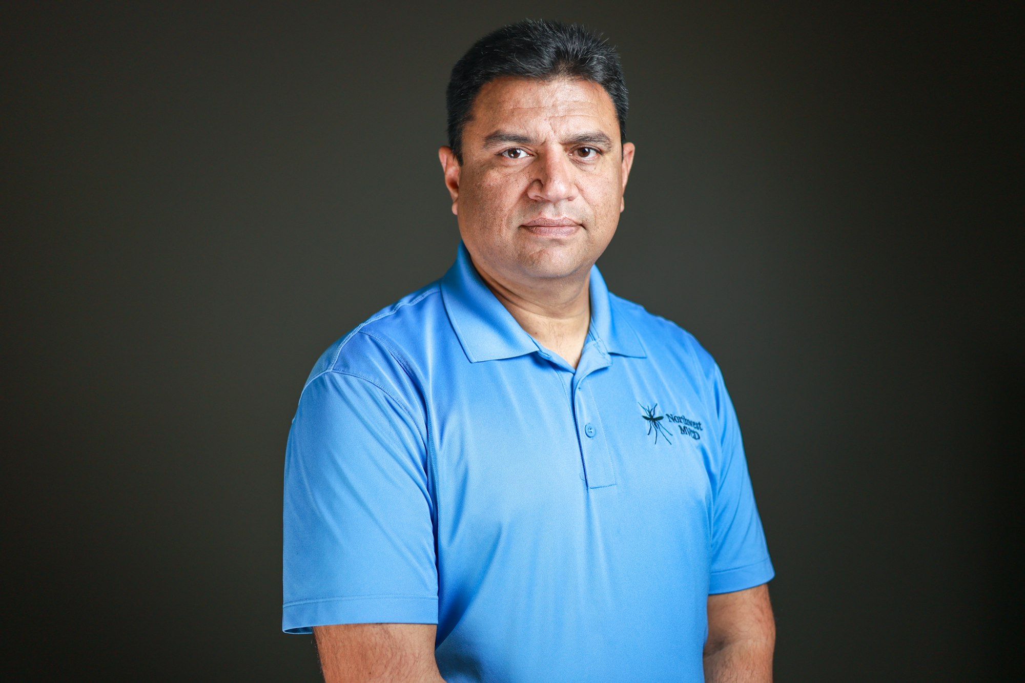 A man wearing a blue polo shirt with a logo stands against a dark background, looking directly at the camera.