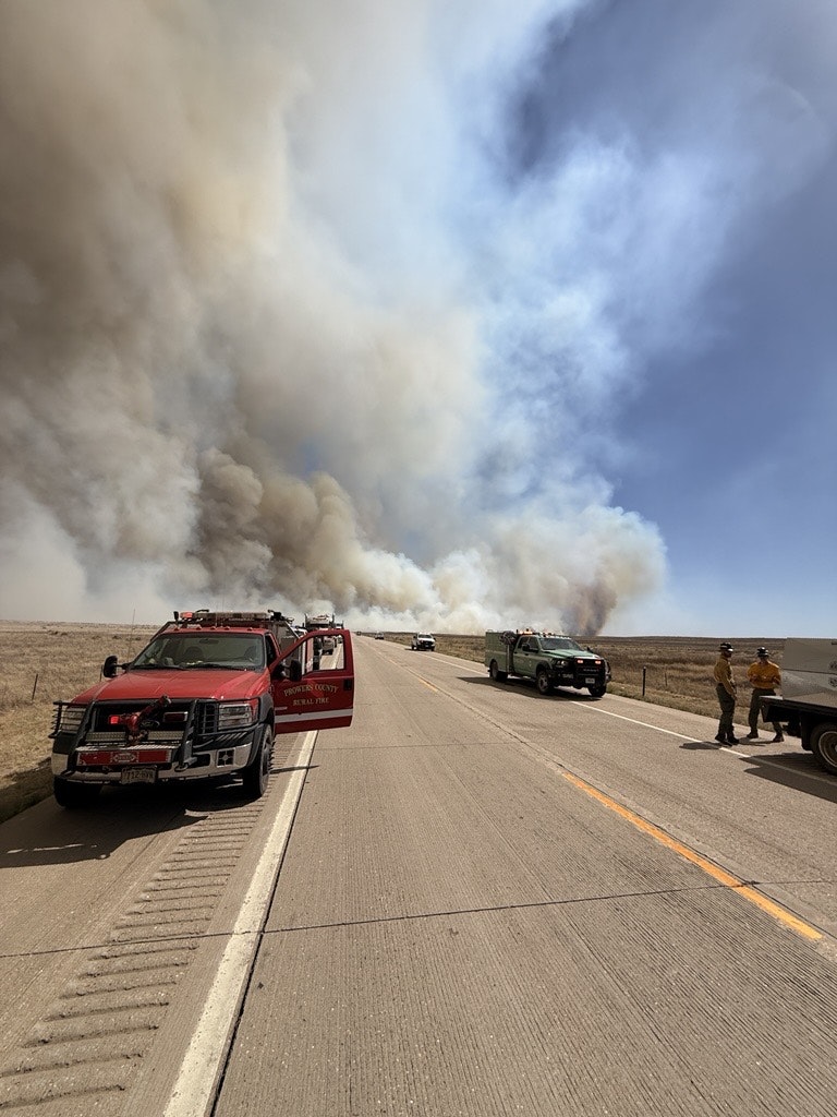 Emergency vehicles line a road amidst heavy smoke from a wildfire in the distance. Firefighters are present, assessing the situation.