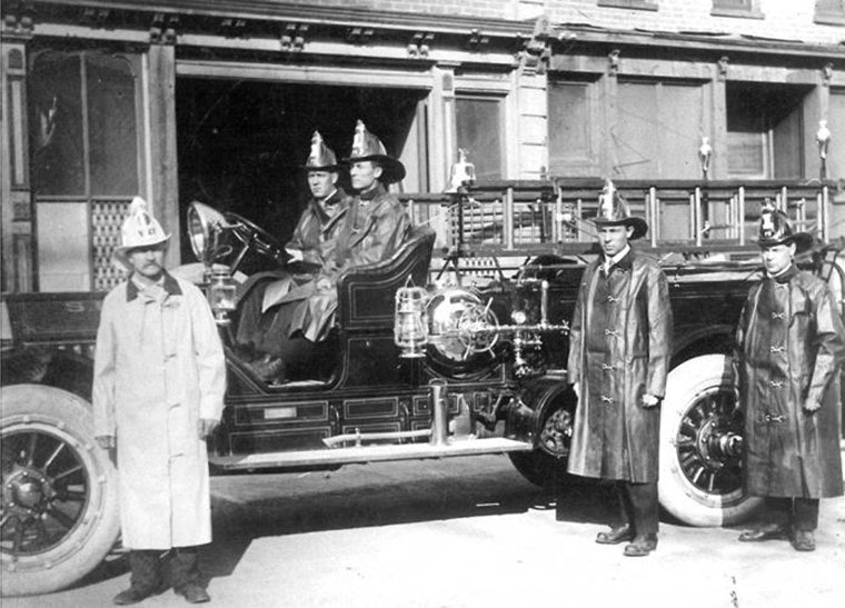 Vintage fire engine with firefighters in uniform and helmets, parked in front of a building.