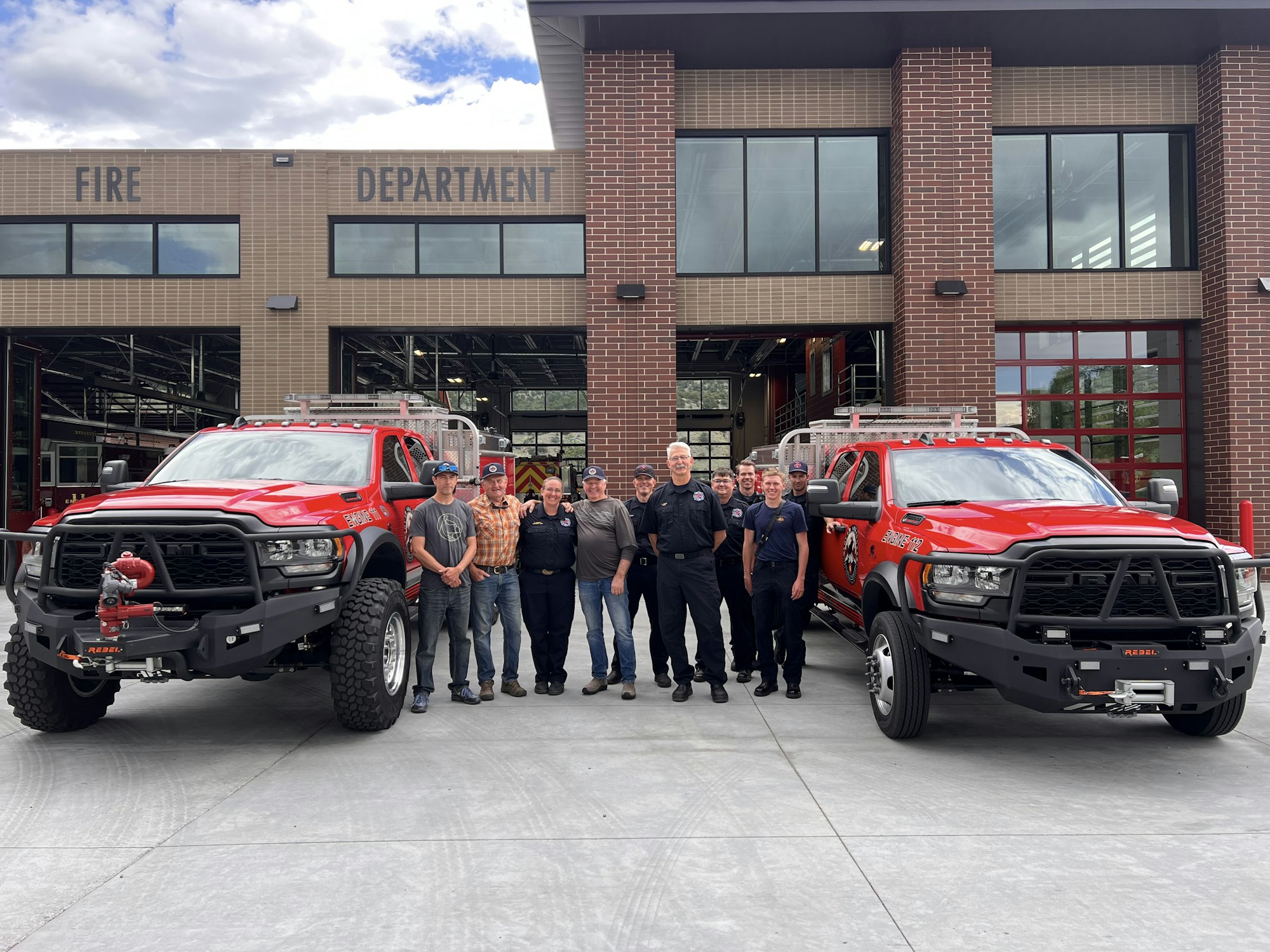 A group of people stand in front of two red fire trucks outside a fire department building.