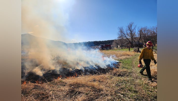 Firefighter conducting controlled burn in field; smoke rising, fire vehicle in the background.