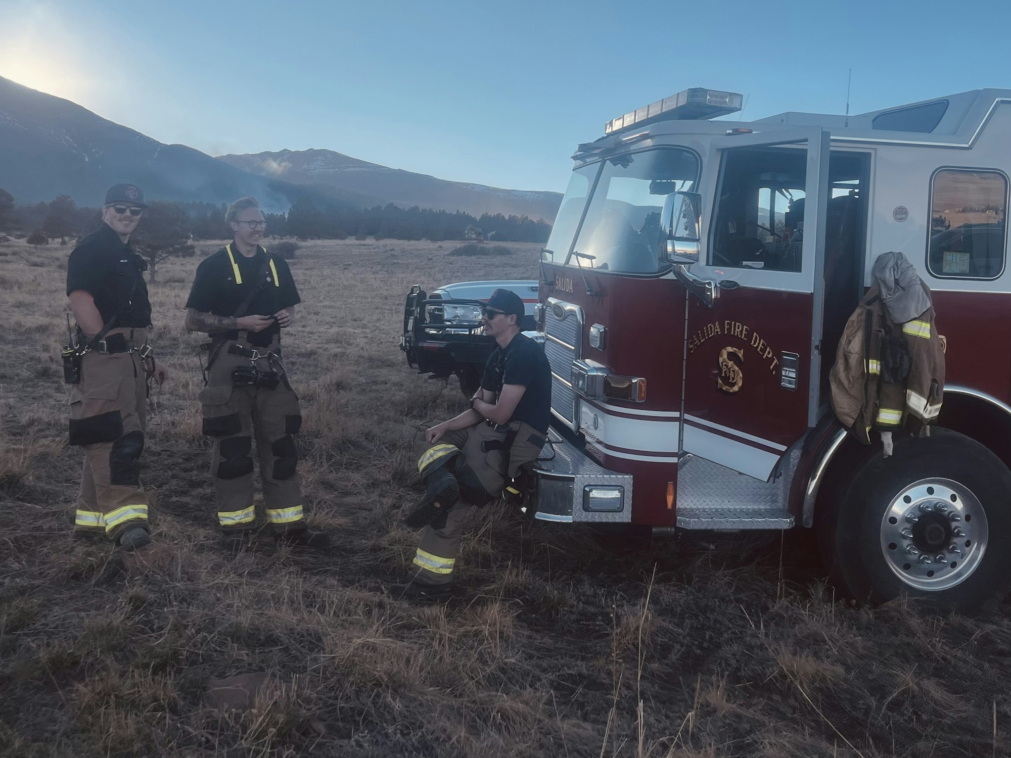 Three firefighters in gear relax by a fire truck in a grassy area, with mountains in the background during sunset.