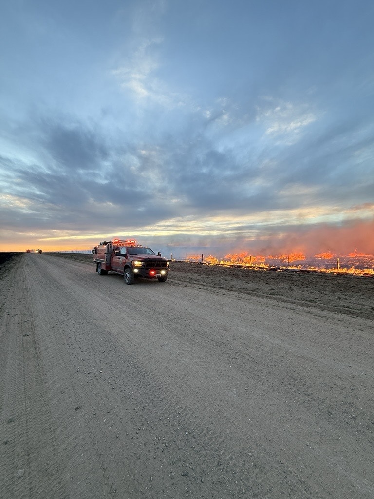 A fire truck on a dirt road near a fire, with dramatic skies and smoke in the background, likely responding to an incident.