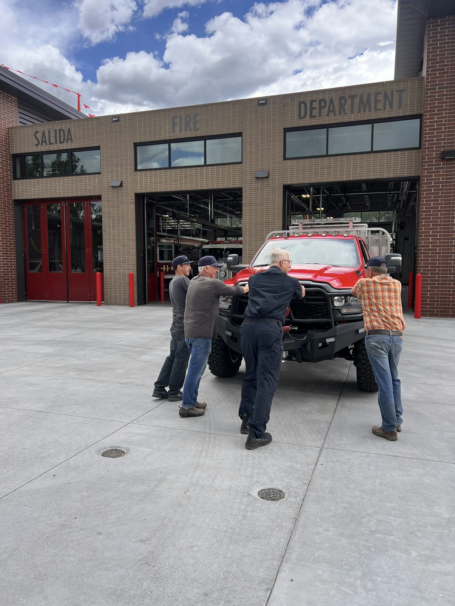 Four people inspect a red vehicle in front of a fire department building.