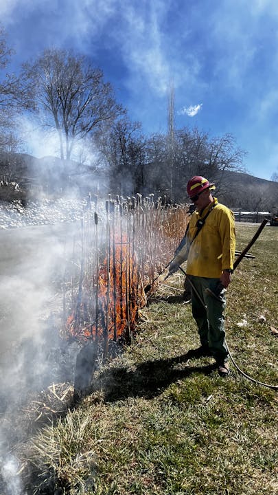A firefighter is managing a controlled burn of dry vegetation under a clear blue sky.
