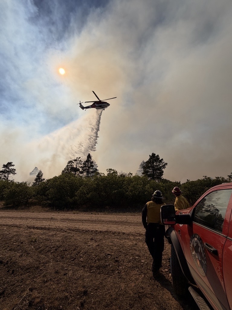 A helicopter drops water on a wildfire as firefighters monitor from the ground under smoky skies.