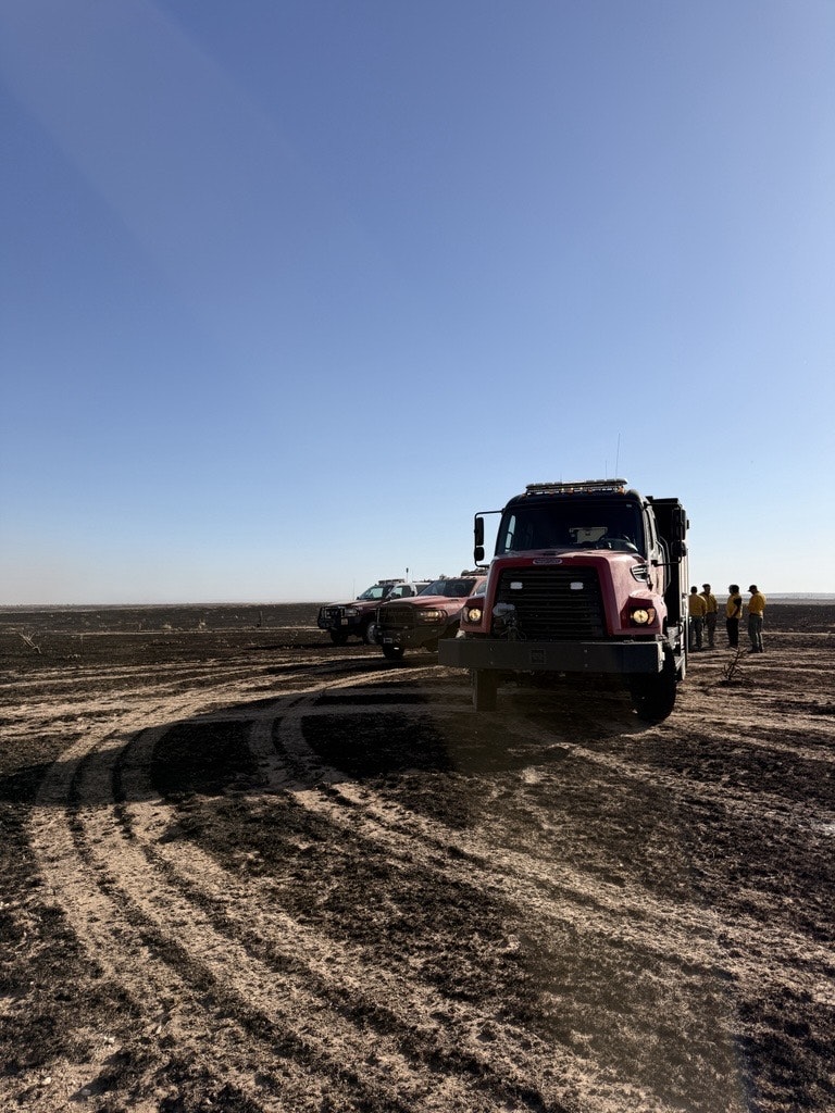 The image shows vehicles in a barren landscape with a clear blue sky and a group of people in yellow shirts nearby.