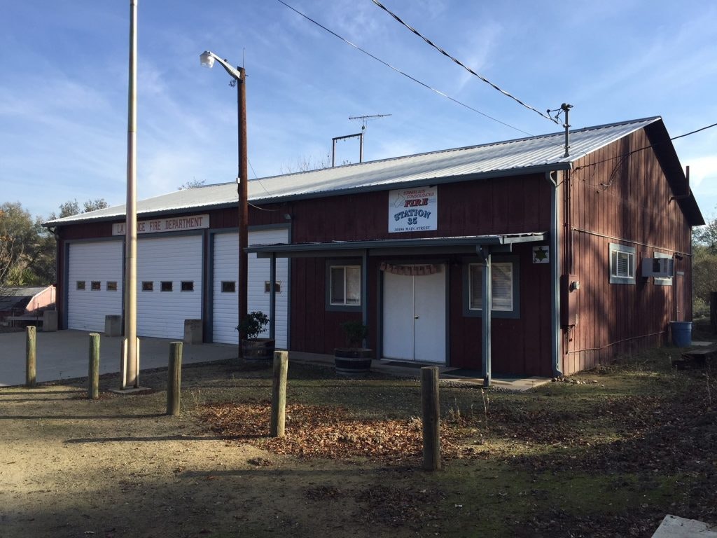 Rustic fire department building with two garage doors, a sign, and surrounding foliage under a clear sky.