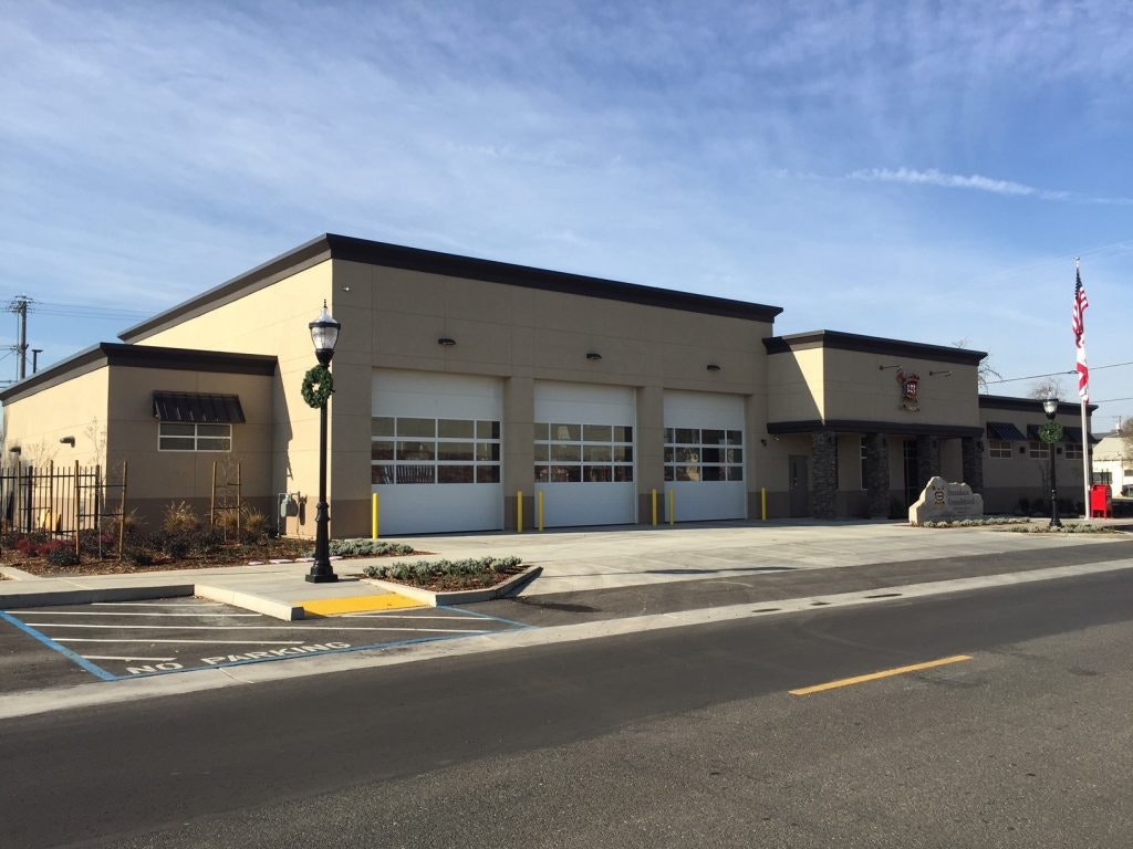 A modern fire station with three garage doors, a flagpole, and clear skies.