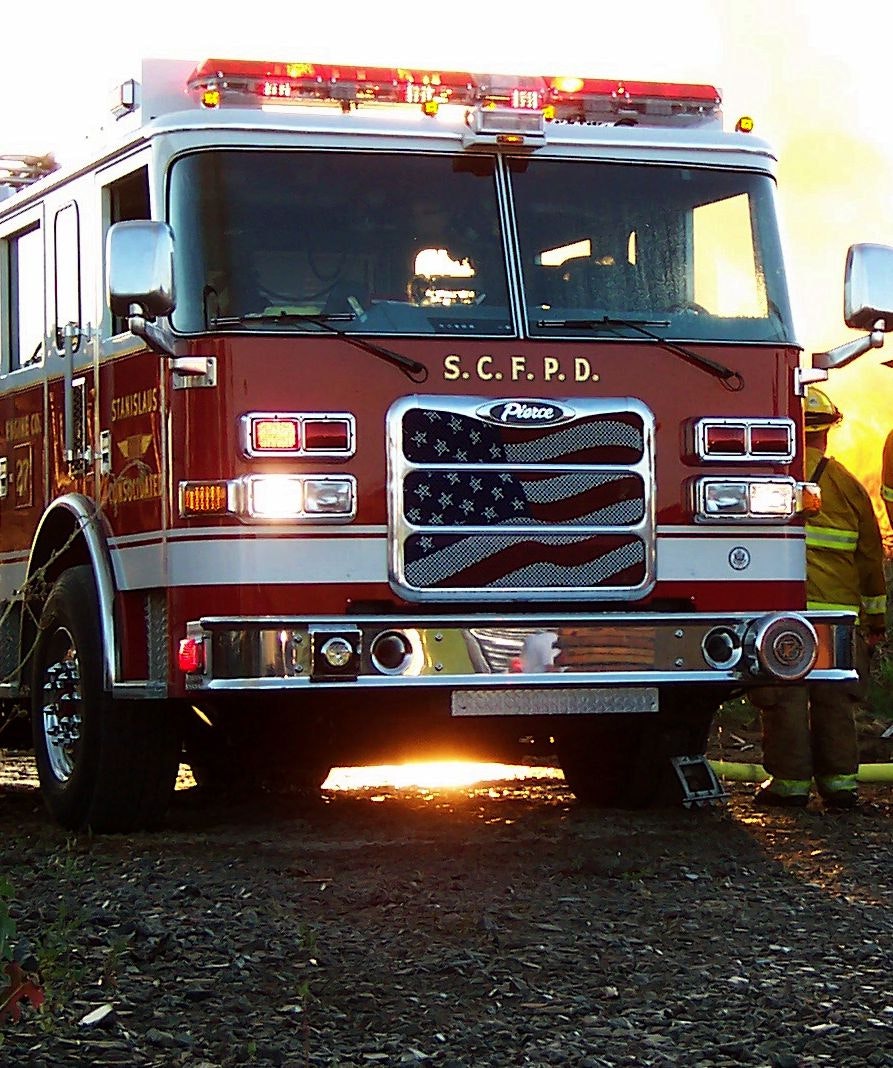 Fire truck with an American flag design on the grill, emergency lights on, firefighter partially visible in the background.