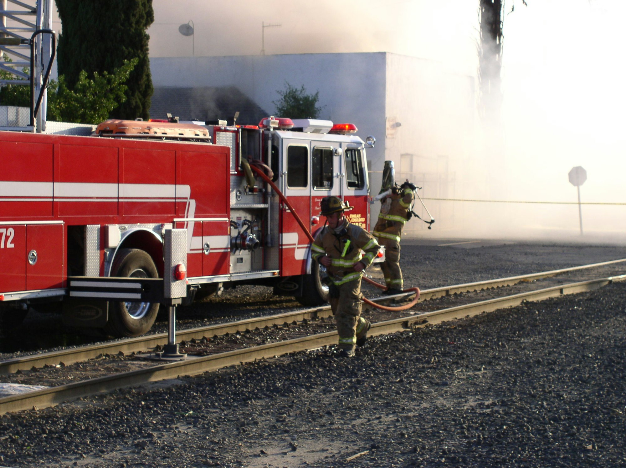 Firefighters with hose near fire truck, smoke billowing from building, near train tracks.