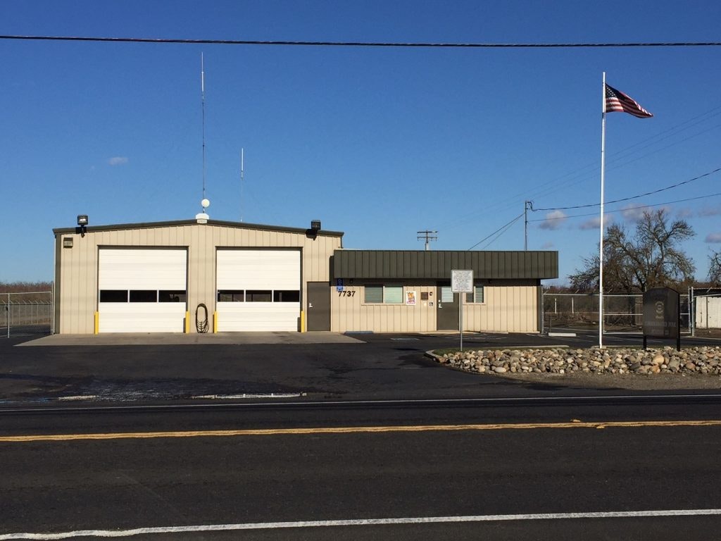 A single-story building with two garage doors, an adjacent flagpole flying the American flag, a clear sky, and a street in front.