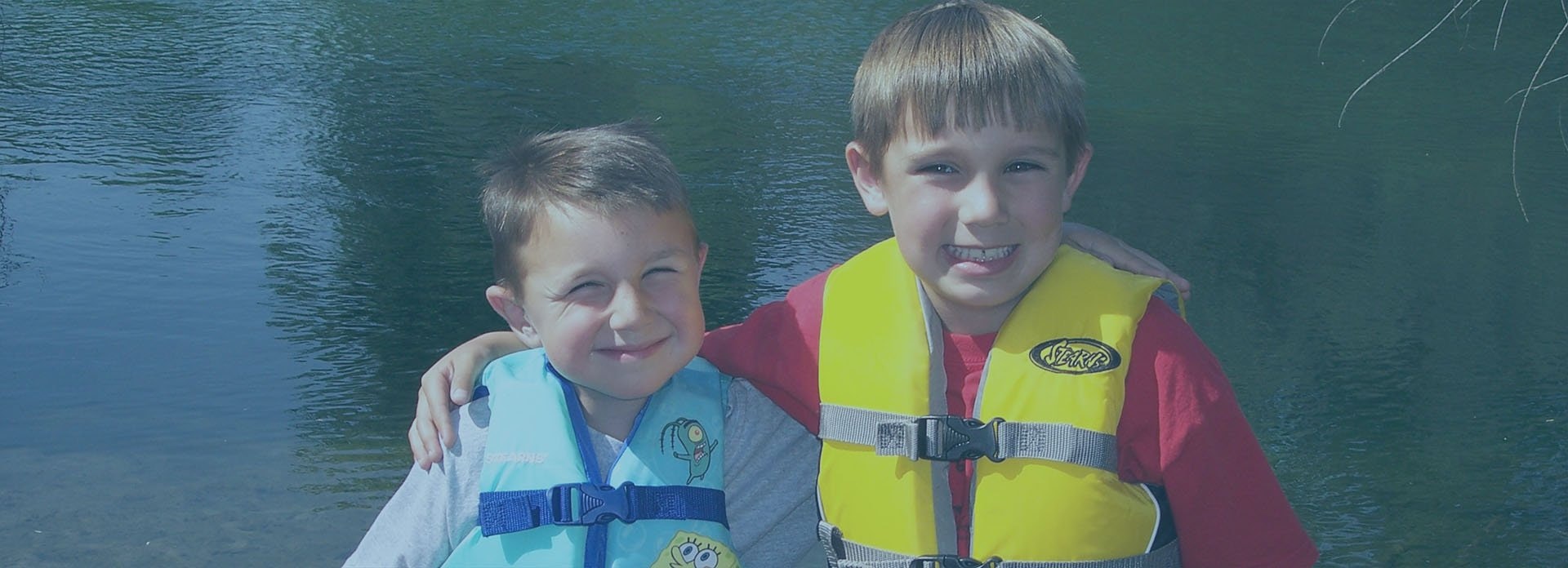 Two smiling kids in life jackets by water.